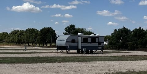 A travel trailer RV parked in a gravel site with green trees in the background.